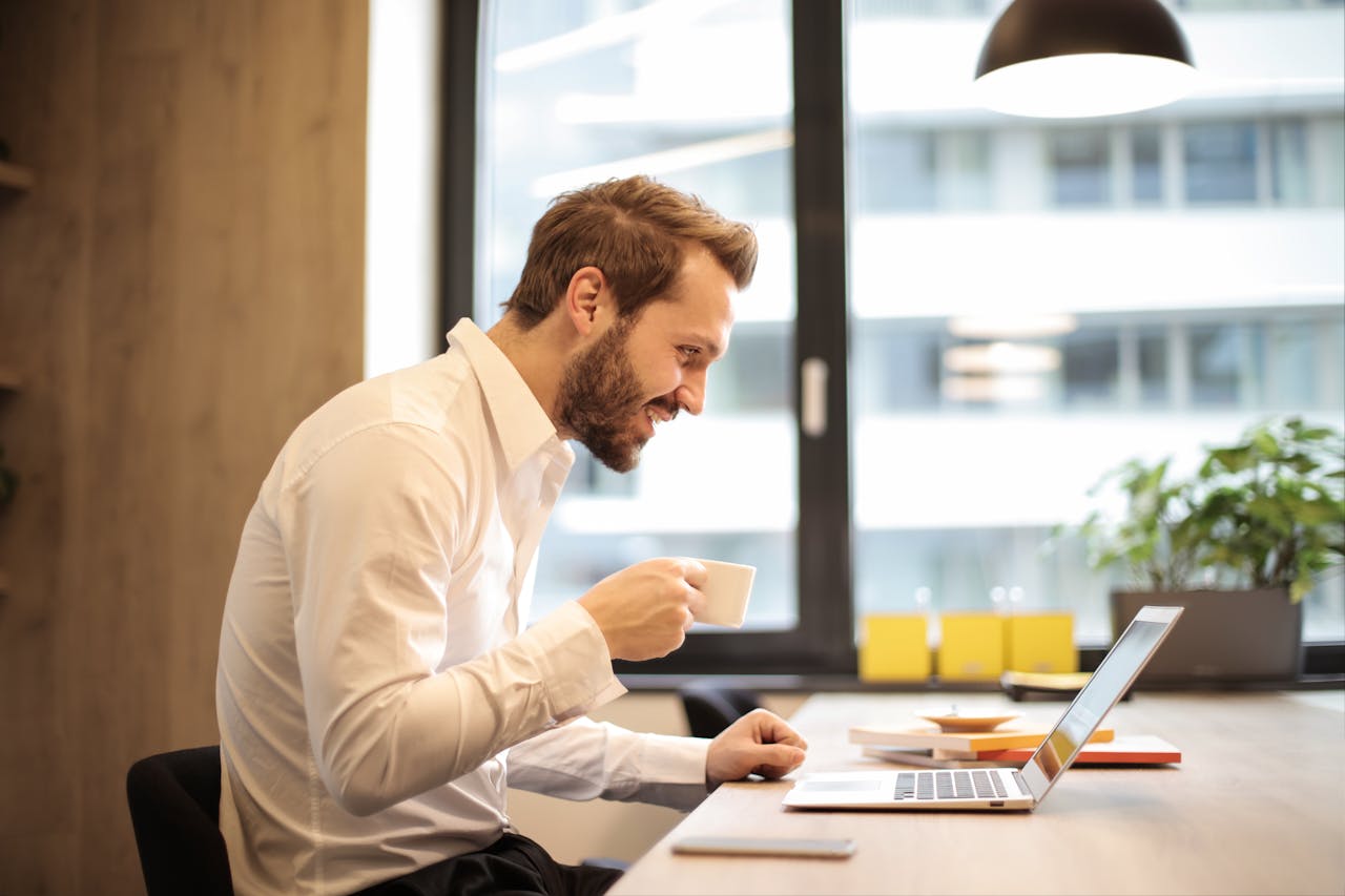 services-bg Man in office with coffee, smiling while working at laptop, captures the essence of remote work.